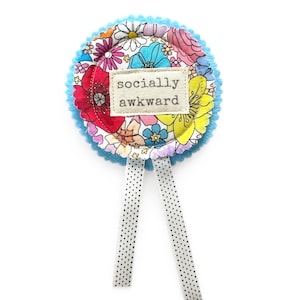 Puede incluir: Una insignia redonda de tela floral con un borde festoneado azul claro y dos cintas blancas con lunares negros. La insignia presenta el texto "socialmente torpe" en una etiqueta rectangular.