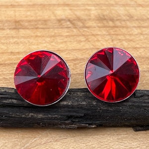 May include: A pair of round stud earrings featuring vibrant red faceted crystals set in a simple silver-toned metal frame. The circular earrings are positioned side by side, showcasing a brilliant, reflective surface with a pointed center design.