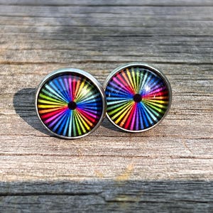 Puede incluir: Un par de pendientes de botón redondos con un diseño vibrante y colorido. Cada pendiente presenta un bisel plateado que rodea una imagen circular con un arcoíris de líneas radiantes, creando un efecto visual llamativo. Los pendientes descansan sobre una superficie de madera envejecida.