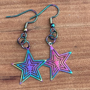 Puede incluir: Un par de pendientes iridiscentes en forma de estrella. Cada pendiente presenta un diseño de estrella en capas con un degradado de colores, incluyendo morado, azul y verde. Los pendientes tienen un estilo de gancho y están conectados por pequeños eslabones circulares.