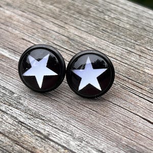 May include: A pair of round stud earrings featuring a black metal setting and a glossy domed surface. Each earring displays a bold, white five-pointed star graphic centered against a solid black background, creating a high-contrast design.