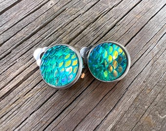 May include: A pair of round clip-on earrings featuring a vibrant, iridescent green and gold scale pattern under a clear domed surface, set within a simple silver-toned metal frame. The design mimics a textured fish or mermaid scale motif.