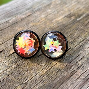 May include: Two round stud earrings with black settings. Each earring features a clear, domed top filled with colorful star-shaped confetti. The stars are in shades of red, yellow, blue, and green, creating a playful and festive look.