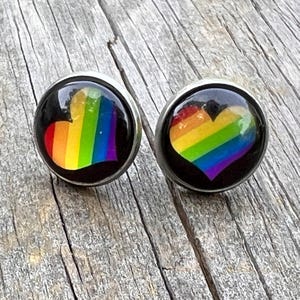 Puede incluir: Un par de pendientes de botón redondos con un diseño de corazón arcoíris. El corazón presenta los colores del arcoíris sobre un fondo negro. Los pendientes tienen un engaste de metal plateado.