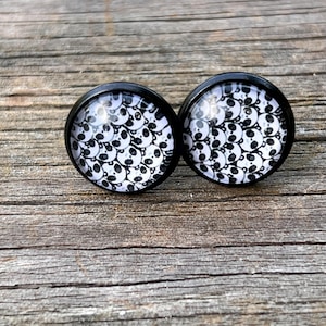 May include: A pair of round stud earrings featuring a black metal frame and a glass cabochon top. The surface of each earring displays a repeating black and white graphic pattern of multiple panda bear faces arranged in a dense, overlapping cluster.