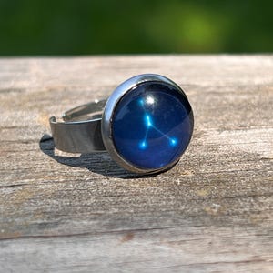 May include: A silver-toned adjustable ring with a round, dark blue cabochon. The cabochon features a constellation design in light blue. The ring is set against a weathered wooden surface with a blurred green background.
