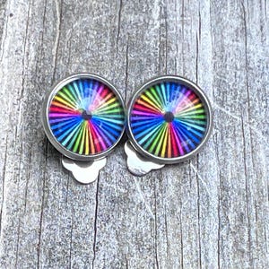 May include: A pair of round, silver-coloured clip-on earrings. Each earring has a vibrant, colourful design with rainbow-coloured stripes radiating from a central black dot. The earrings are set against a weathered, grey wooden surface.