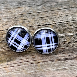 May include: A pair of round stud earrings featuring a black, white, and light purple plaid pattern under a clear glass dome. The circular designs are set within simple silver-toned metal frames, creating a clean and symmetrical appearance.