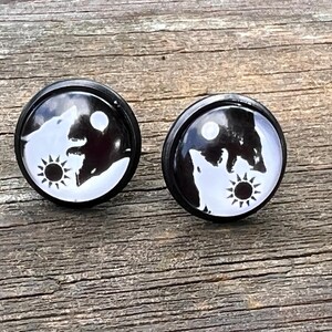 May include: A pair of round stud earrings featuring a black and white graphic design. Each earring displays a silhouette of a wolf howling at a moon, accompanied by a sun icon, arranged in a circular composition with a smooth, glossy finish.