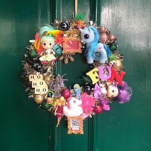 以下が含まれることがあります： 人形、おもちゃの車、マイリトルポニーのフィギュアなど、おもちゃやオーナメントが飾られた気まぐれなリース。リースは金色のきらびやかなもので、「FUN」の文字がカラフルに書かれています。ドア番号「35」が上にあります。