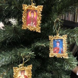 May include: Three Christmas tree decorations featuring images of David Bowie in ornate gold frames. The frames are rectangular with a decorative, glittery border. The ornaments hang from gold-coloured strings, set against a green Christmas tree.