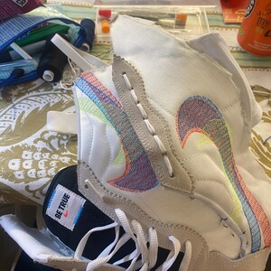 May include: A white and black sneaker with colorful stitching and the words "Be True" on the side. The sneaker is being repaired with a needle and thread.