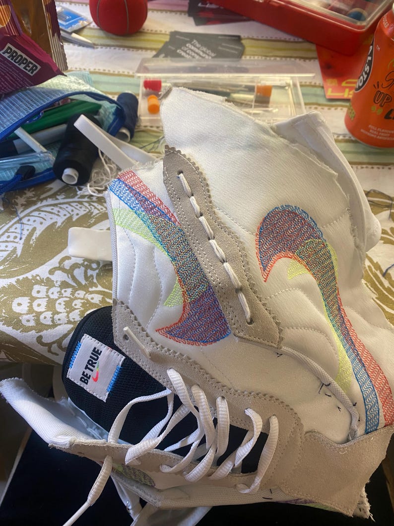 May include: A white and black sneaker with colorful stitching and the words "Be True" on the side. The sneaker is being repaired with a needle and thread.