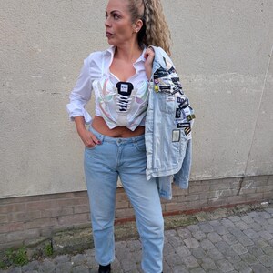 May include: A woman wearing a white shirt, light blue jeans, and black ankle boots. She is wearing a white corset with a sneaker design. She is holding a light blue denim jacket with patches on it.