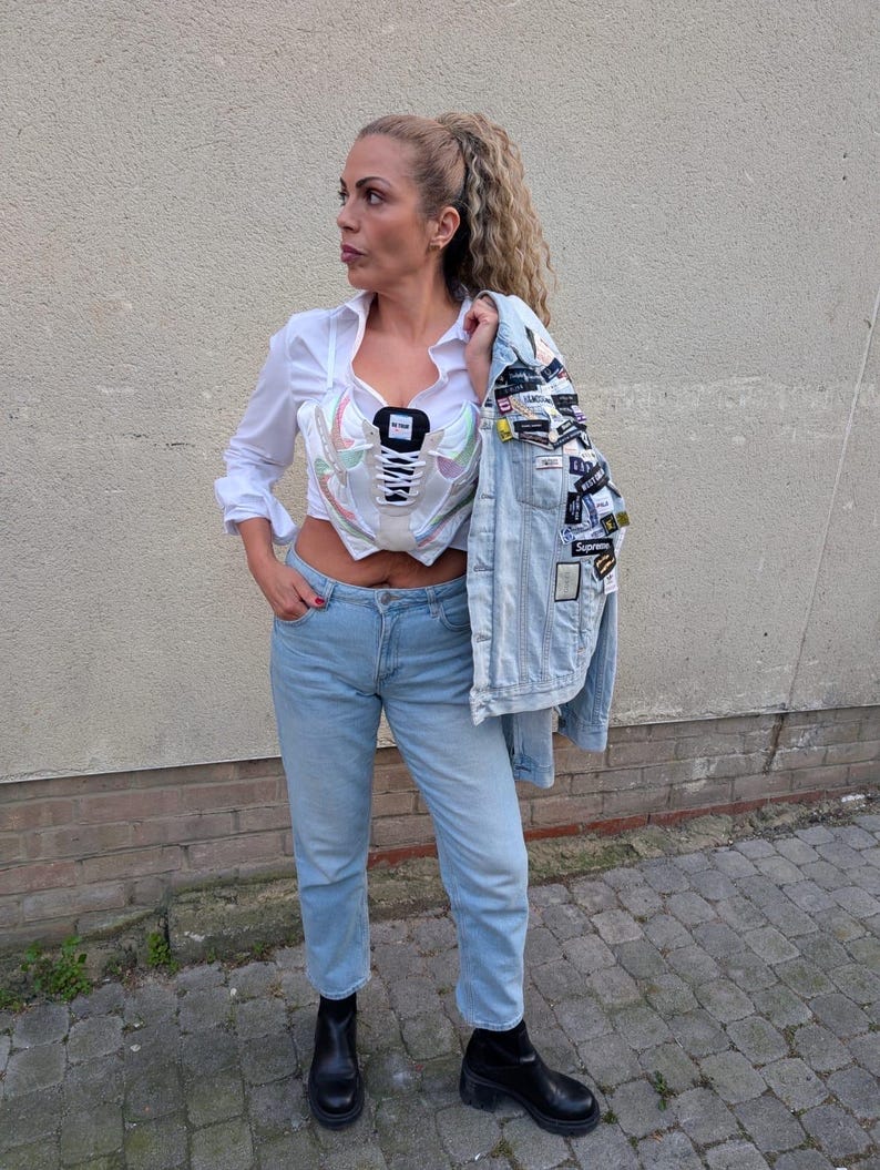 May include: A woman wearing a white shirt, light blue jeans, and black ankle boots. She is wearing a white corset with a sneaker design. She is holding a light blue denim jacket with patches on it.