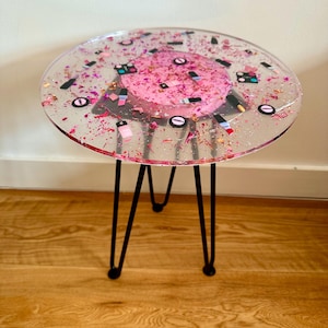 May include: A round side table with a clear resin top. The top is decorated with pink glitter, small nail polish bottle shapes, and a central pink circle. The table has black hairpin legs.