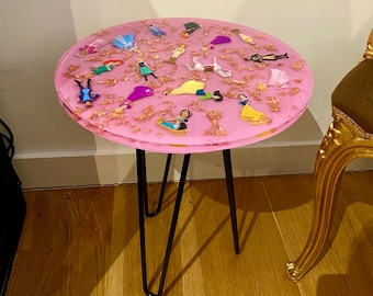 Puede incluir: Una mesa redonda rosa con una tapa de resina que presenta una variedad de figuritas de princesas Disney incrustadas en la resina. La mesa tiene patas de horquilla negras.