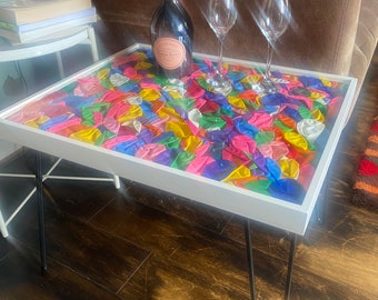 May include: A white coffee table with black hairpin legs. The table top is made of clear resin with colorful balloons embedded inside. Two wine glasses and a bottle of champagne are on the table.