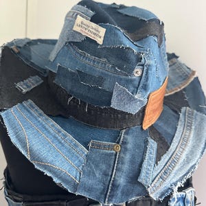 May include: A patchwork denim cowboy hat with a brown leather band and a black band around the crown. The hat is made from various shades of blue denim and features a Levi's Premium label.