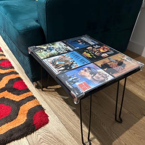 May include: A black side table with a glass top featuring a collage of DVD cases. The table has four thin black metal legs and is sitting on a wooden floor with a red and orange patterned rug.