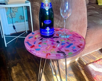 Puede incluir: Una botella de champán Pommery y una copa de vino descansan sobre una pequeña mesa redonda rosa. La mesa tiene una tapa de resina transparente con decoraciones de zapatos en miniatura de colores y patas de horquilla de metal.