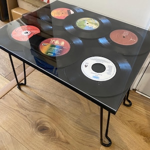 Könnte beinhalten: Ein schwarzer Couchtisch mit einer Glasplatte und einem Design aus Vintage-Schallplatten. Der Tisch hat vier schwarze Metall-Haarnadelbeine.