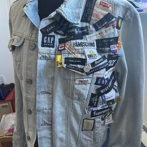 May include: Pale blue denim jacket embellished with a collage of brand labels, such as Gap, Tommy Hilfiger, and Gucci. The jacket has a traditional button-up style, a collared neckline, and pockets. The labels are diverse in size and colour, creating a distinctive, patchwork appearance.