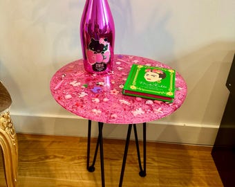 Puede incluir: Una pequeña mesa auxiliar redonda rosa con patas de horquilla negras. La superficie de la mesa está decorada con pequeños adornos blancos y rosas. Una botella rosa con una etiqueta de Hello Kitty y un libro verde están sobre la mesa. Un arreglo de plumas rosas está en la botella.