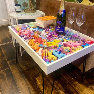 May include: A white coffee table with black hairpin legs. The table top is filled with colourful plastic toy horses. A bottle of champagne and two champagne glasses are on the table.