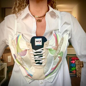 May include: A white shirt with a pair of white sneakers worn as a top. The sneakers have rainbow colored accents and the text "BE TRUE" on the tongue.