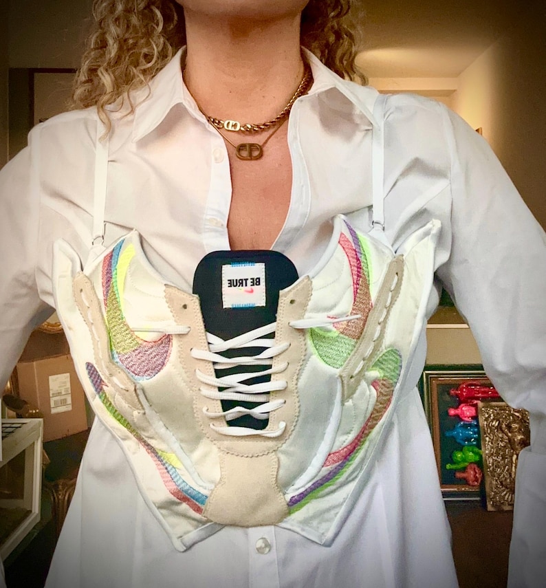May include: A white shirt with a pair of white sneakers worn as a top. The sneakers have rainbow colored accents and the text "BE TRUE" on the tongue.