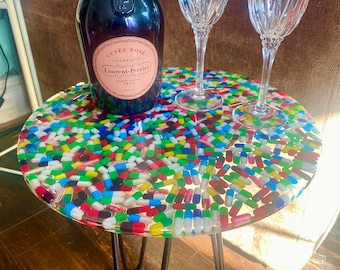 May include: A round side table with a colorful pill design on the tabletop. The table has black hairpin legs. A bottle of Cuvée Rosé Champagne and two crystal wine glasses are on the table.
