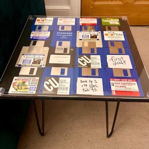 May include: A square coffee table with a clear glass top, featuring a collage of vintage 3.5-inch floppy disks. The disks are blue, black, and white, with labels and text. The table has black hairpin legs.