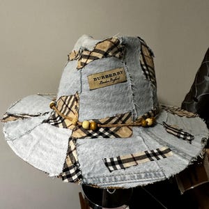 May include: A patchwork denim hat with a wide brim. The hat features patches of Burberry plaid fabric and a leather tag that reads "BURBERRY London, England". A string with wooden beads decorates the hat.