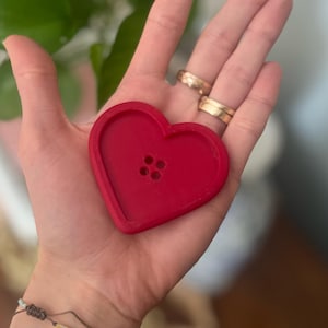 Puede incluir: Un botón rojo brillante en forma de corazón con tres pequeños agujeros. El botón se sostiene en una mano, con anillos de oro en los dedos y varias pulseras en la muñeca. El fondo está borroso, con hojas verdes visibles.