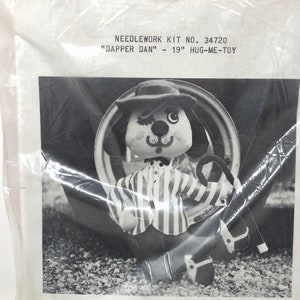 May include: A vintage needlework kit featuring a plush dog toy named "Dapper Dan." The image shows the toy in a striped shirt, hat, and cane. The kit is labeled as 19 inches in size.