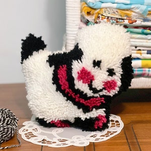 Pu&ograve; includere: Un cane di peluche bianco e nero con accenti rosa. Il cane &egrave; seduto su un pizzo bianco su una superficie di legno.