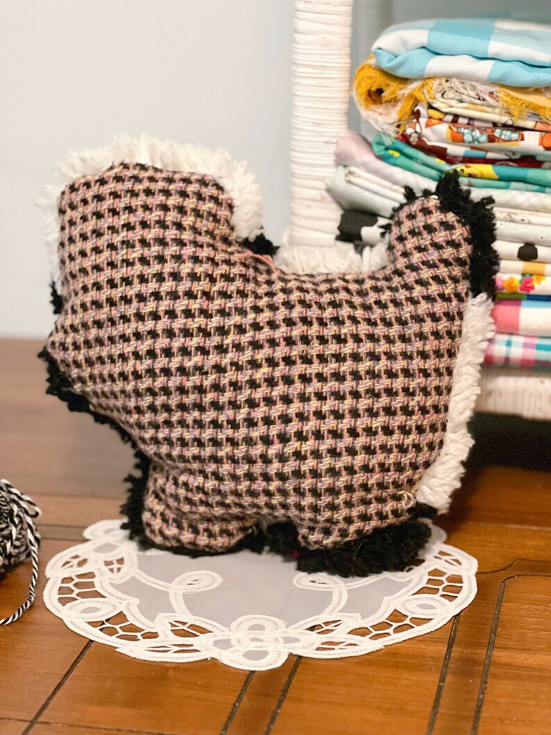 Pu&ograve; includere: Un peluche a forma di cane, a scacchi neri e rosa. Il cane ha la pelliccia bianca intorno al viso e ai piedi. Il cane &egrave; seduto su un pizzo bianco su un pavimento di legno.