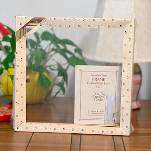 Può includere: Una cornice per foto dorata con un passepartout bianco e un bordo con un motivo blu e bianco. La cornice è confezionata in un sacchetto di plastica trasparente e include un piccolo pezzo di carta con il testo "Another Fine FRAME Crafted With Love By: The Creative Circle 5208 9" x 9" Antique Gold".
