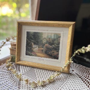 Op de afbeelding: Een ingelijste prent met een goudkleurige lijst. De prent toont een huisjesscène met een witte poort en groen. De tekst "Live in a Romantic Light" staat boven de afbeelding. De lijst rust op een kanten kleedje.