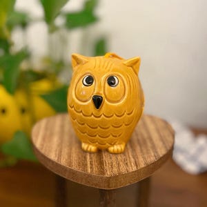 Może przedstawiać: Ceramiczna figurka sowy z żółtą glazurą i czarnymi oczami. Sowa siedzi na drewnianym stojaku w kształcie serca.