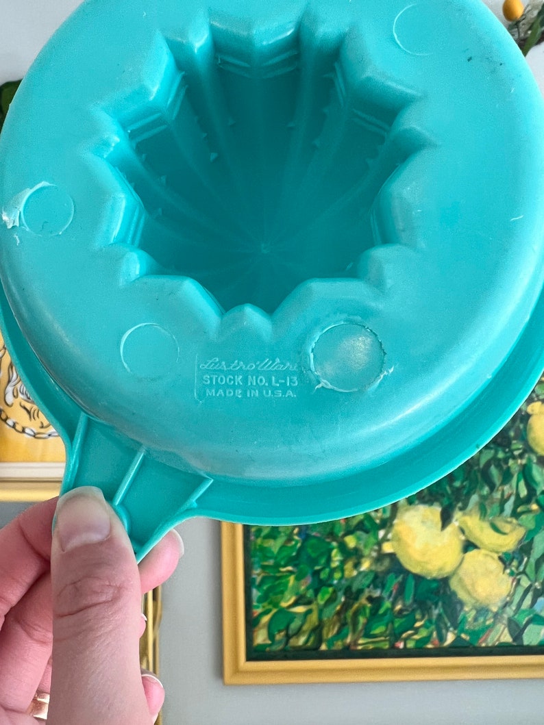 Pode incluir: Um espremedor de citrinos de pl&aacute;stico azul turquesa com uma pega. O espremedor tem um design de estrela e o texto "Dixieware Stock No. L-13 Made in U.S.A." impresso na parte inferior.