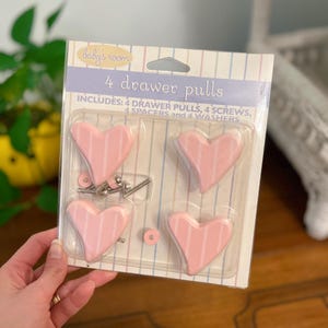 以下が含まれることがあります： 白いストライプの入ったピンクのハート型の引き出しノブが4個入ったパッケージ。パッケージにはネジ、スペーサー、ワッシャーが含まれています。  "Baby's room 4 drawer pulls"とパッケージに印刷されています。