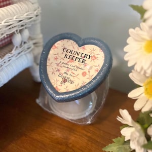 Op de afbeelding: Een hartvormig potdeksel met de tekst "COUNTRY KEEPER" en een bloemmotief. Het deksel is blauw met een bloemmotief en de tekst "A Quality Craft Product Made in USA Mfg. by the New Berlin Co."