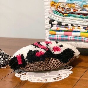 Pu&ograve; includere: Uno scaffale in vimini bianco con una pila di ritagli di stoffa colorati. Un cuscino soffice nero, bianco e rosa si trova su un pizzo bianco su un pavimento di legno.
