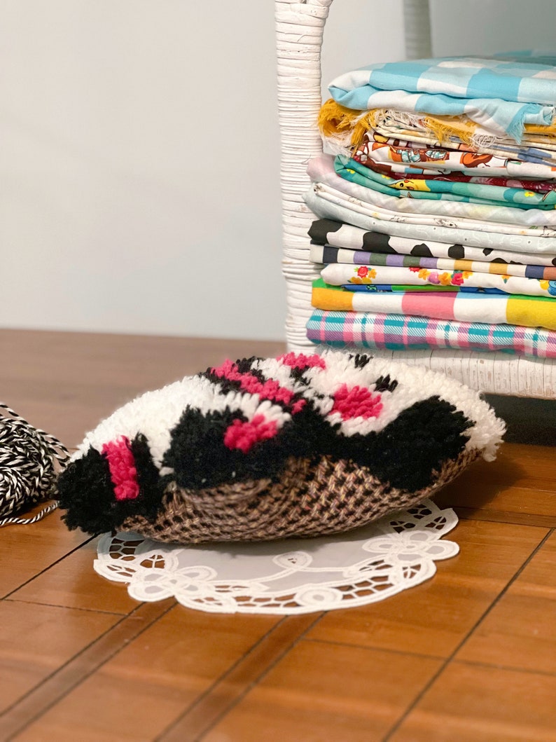 Pu&ograve; includere: Uno scaffale in vimini bianco con una pila di ritagli di stoffa colorati. Un cuscino soffice nero, bianco e rosa si trova su un pizzo bianco su un pavimento di legno.