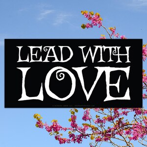 May include: Black and white sign with the text "Lead with Love" in a stylized font. The sign is against a blue sky with a pink flowering tree in the foreground.