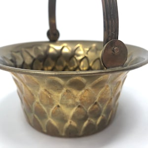 Pode incluir: Uma pequena cesta decorativa de latão com alça. A cesta tem um padrão texturizado em forma de diamante.