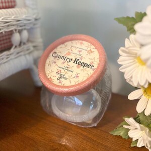 Op de afbeelding: Een heldere glazen pot met een roze deksel met de tekst "Country Keeper" en bloemmotieven. De pot is verpakt in plastic en staat op een houten oppervlak. De deksel heeft een ontwerpopening van 8,57 cm.