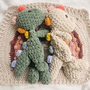 May include: Two crocheted dinosaur plush toys, one in sage green and one in cream, featuring small colorful spikes along their backs. They are positioned on top of a square, crocheted textured mat with a layered, multi-colored rainbow pattern.